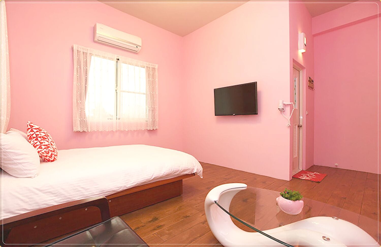 Un dormitorio con decoración rosa, una cama, un televisor, una mesita de cristal y una puerta.