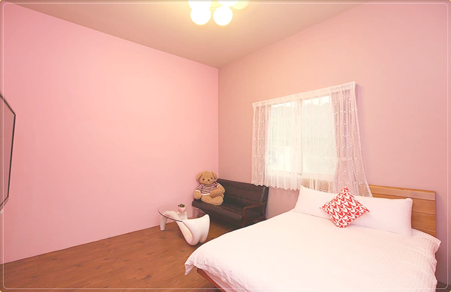 Un dormitorio con cama, una mesita, una silla, un osito de peluche y una ventana con cortinas ligeras.