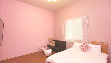 Un dormitorio con cama, una mesita, una silla, un osito de peluche y una ventana con cortinas ligeras.