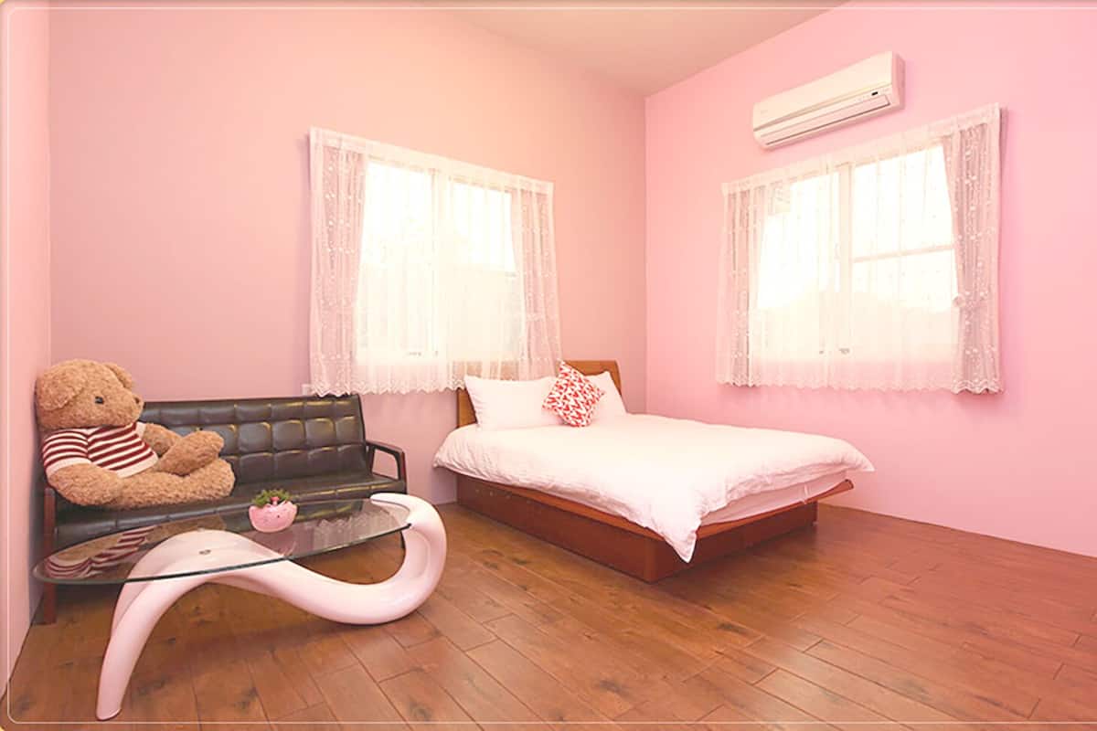 A pink-themed bedroom with a bed, two windows with lace curtains, a teddy bear on a sofa, and a glass coffee table.