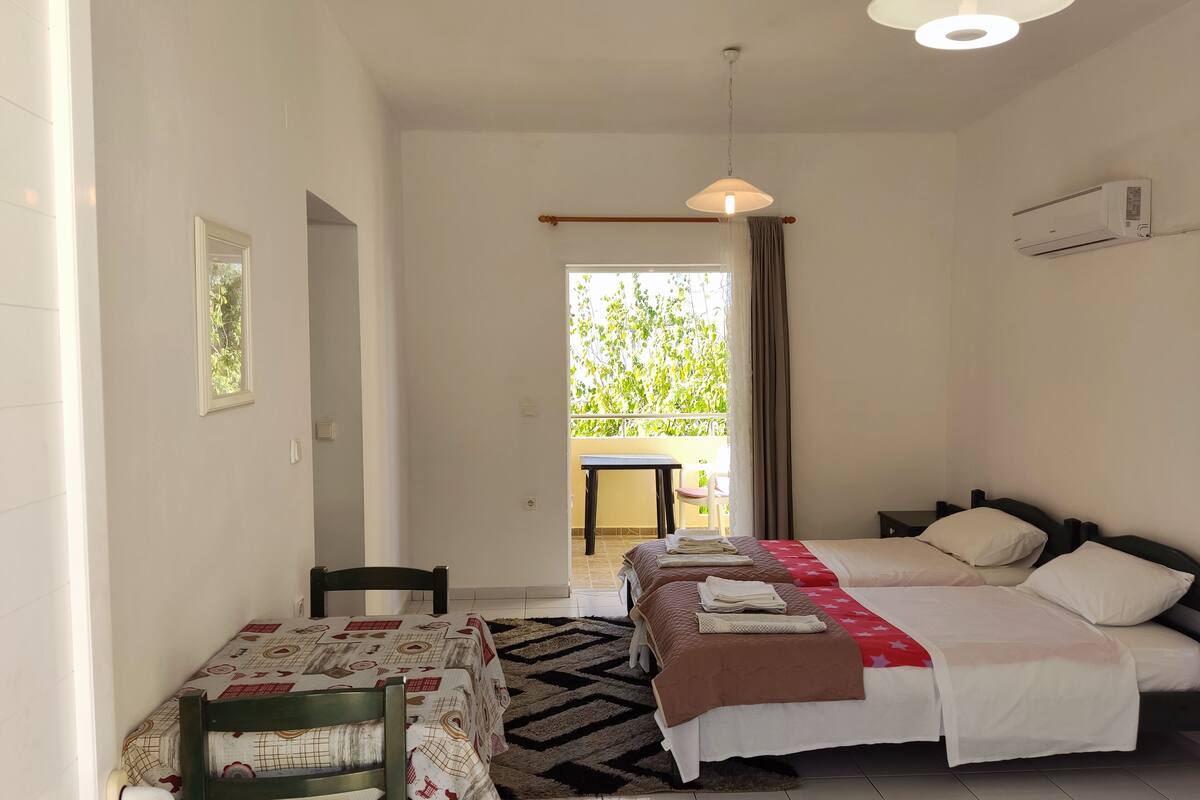 A bedroom with two beds, a table, and a chair. There is a window with curtains, a ceiling light, and an air conditioning unit.