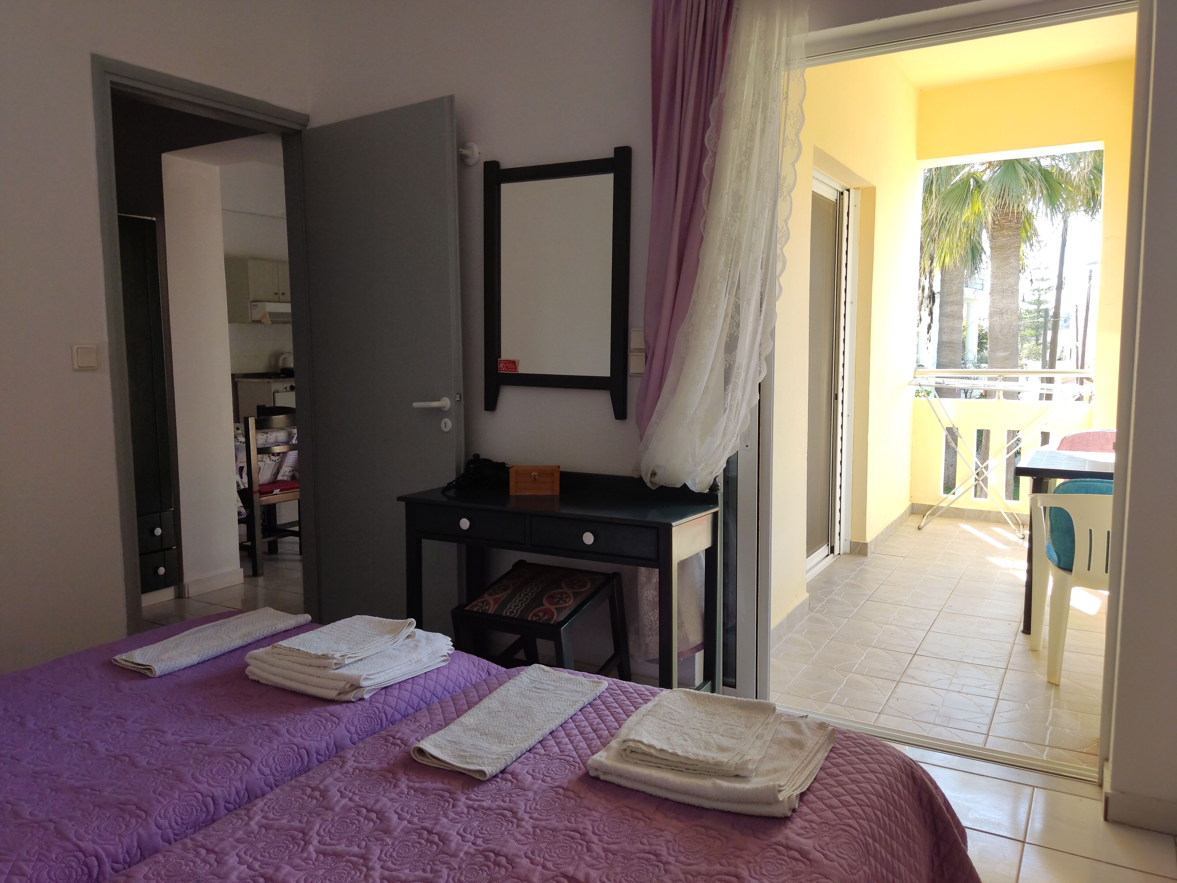 A hotel room with a bed, a desk, a mirror, and a balcony with a view of palm trees.