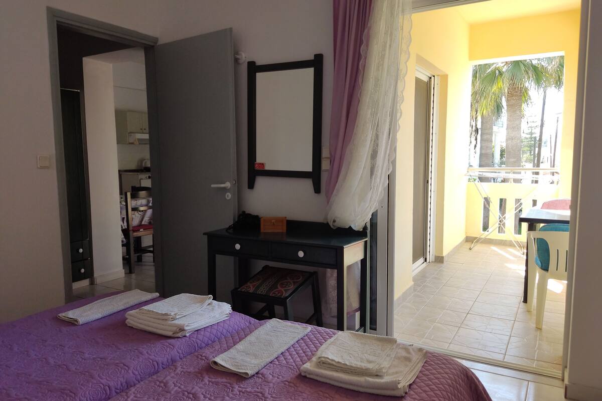 A hotel room with a bed, a desk, a mirror, and a balcony with a view of palm trees.