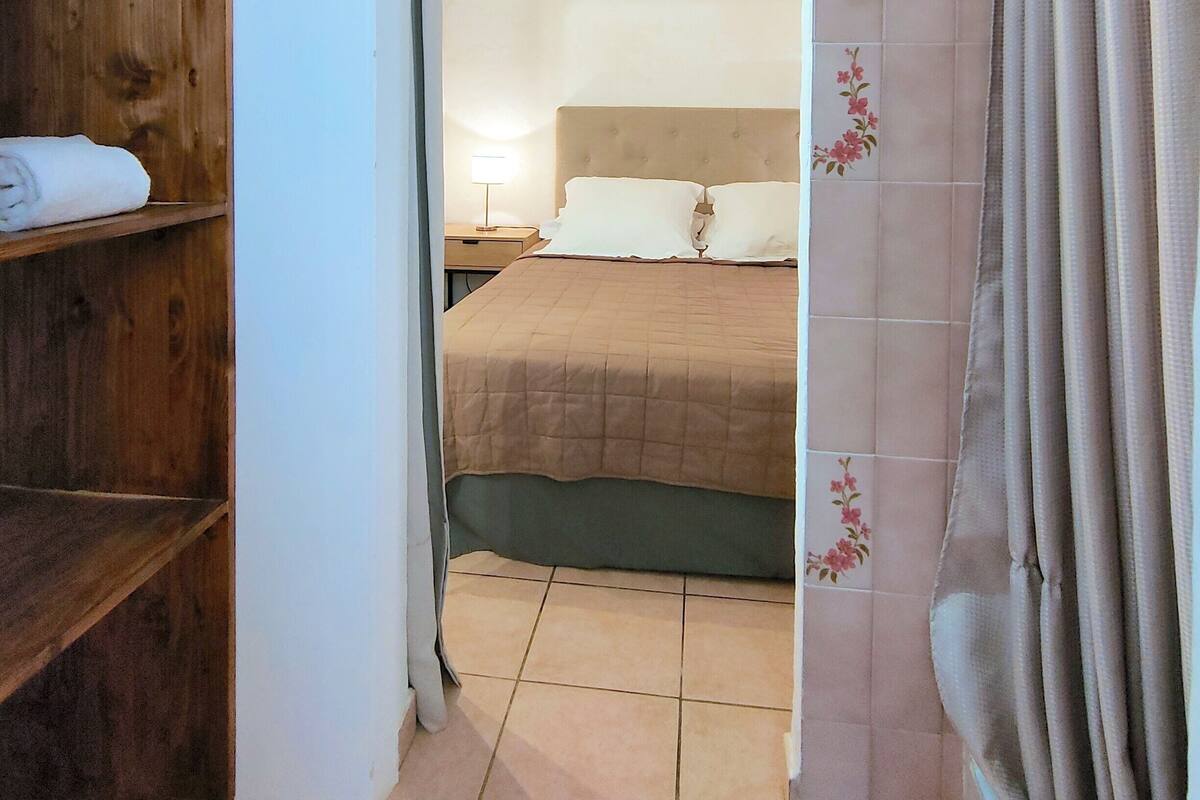 A narrow hallway leading to a bedroom with a bed, a wooden shelf with towels, and a curtain with floral patterns.