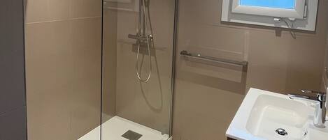 Un baño moderno con ducha de vidrio, lavamanos sobre mueble de madera y ventana sobre el espejo.