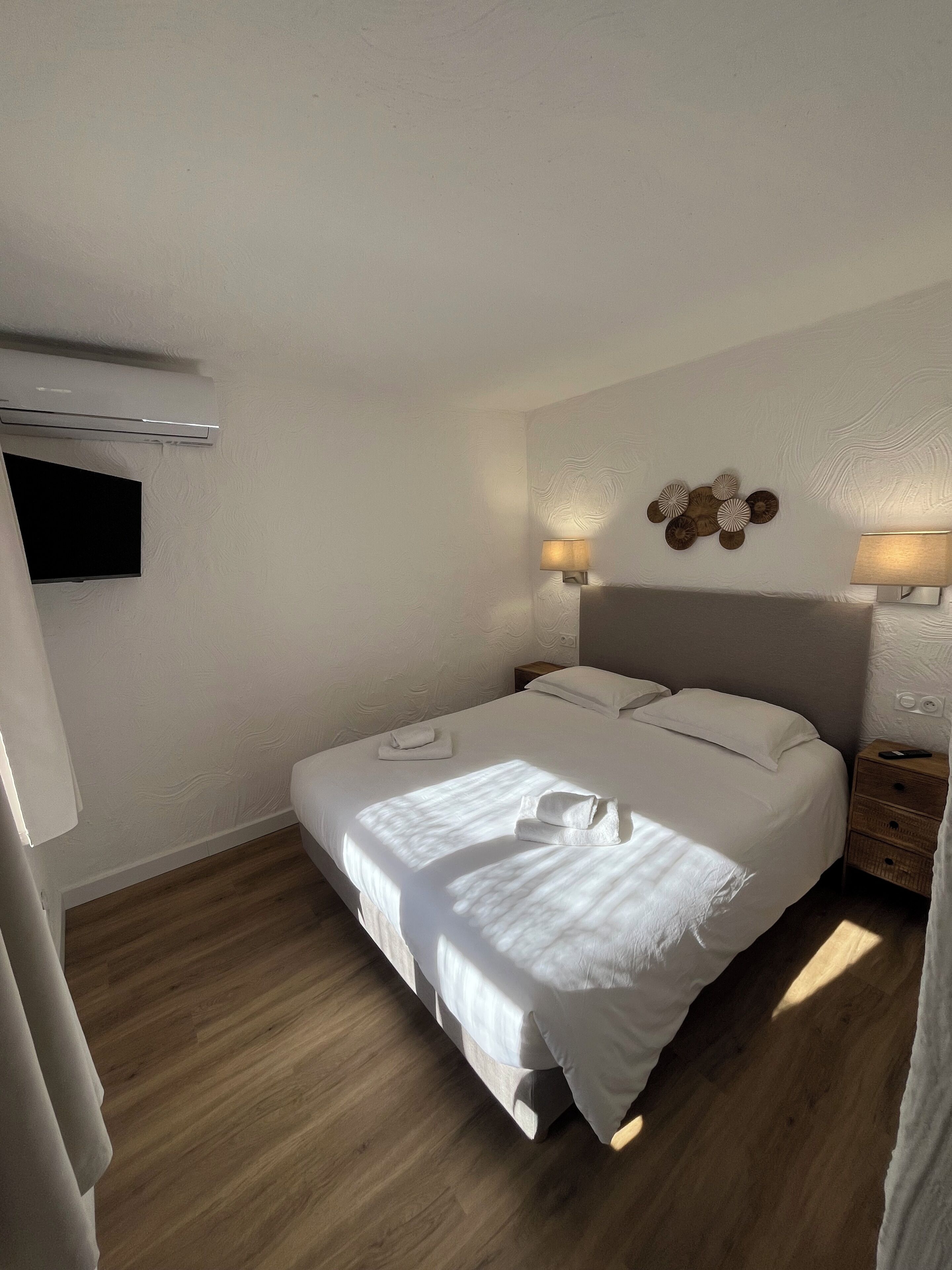 A neatly made bed with white linens, a flat-screen TV mounted on the wall, two bedside lamps, and a wooden nightstand with a remote control.