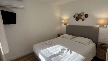 A neatly made bed with white linens, a flat-screen TV mounted on the wall, two bedside lamps, and a wooden nightstand with a remote control.