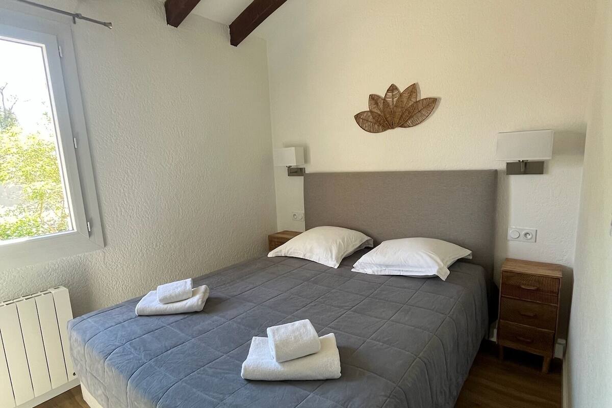 A bedroom with a bed, wooden beams, a radiator, a window, and a wall-mounted lamp.