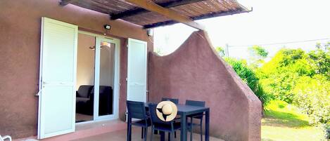 Une terrasse couverte avec une table et des chaises, une porte donnant sur l’intérieur et une vue sur la verdure en arrière-plan.