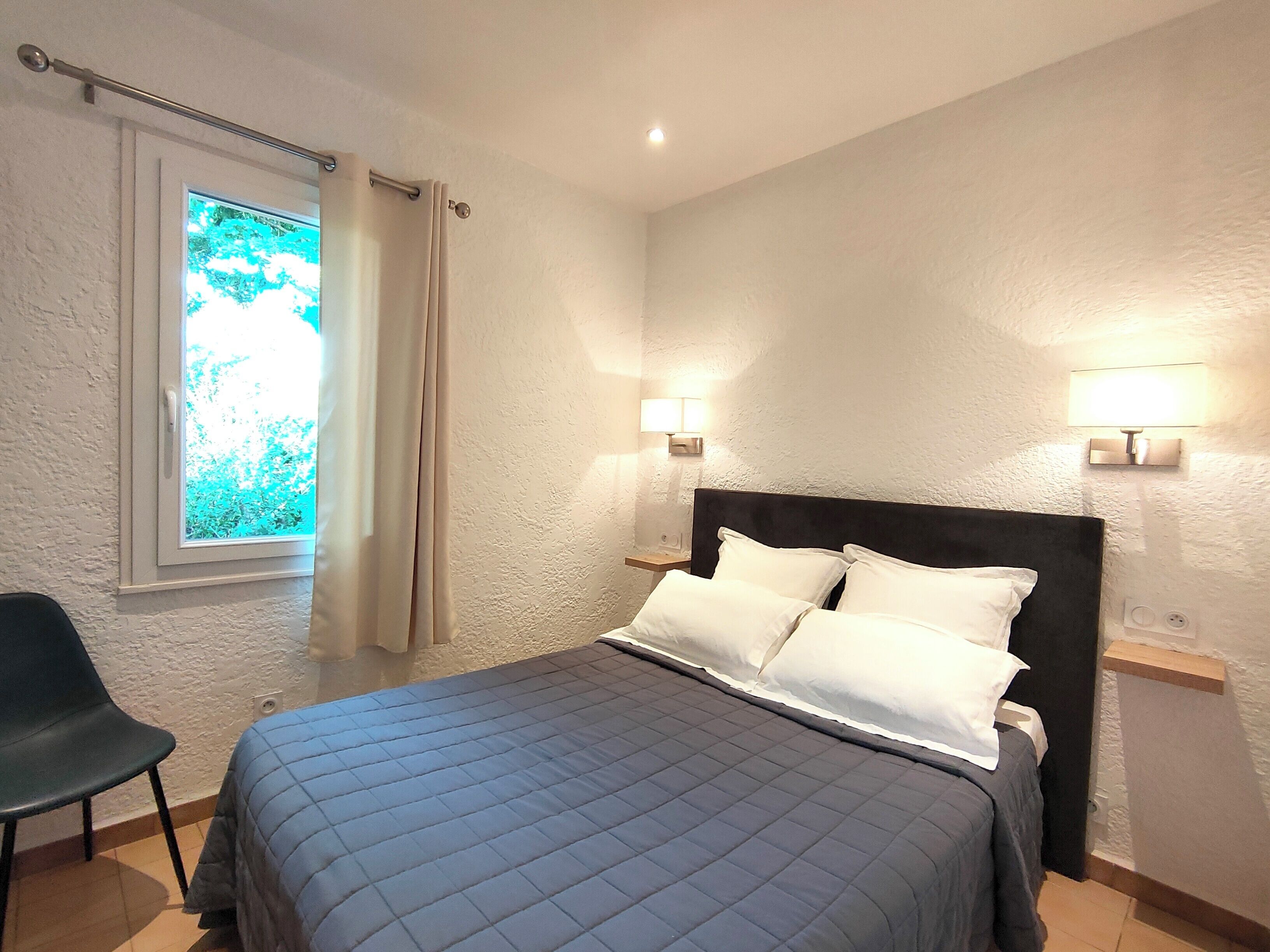 A bedroom with a bed, two bedside lamps, a window with curtains, and a chair.
