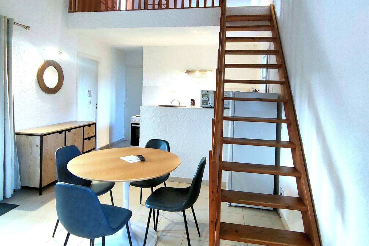 A dining area with a wooden table and blue chairs, a lofted sleeping area with a wooden ladder, and a kitchen in the background.