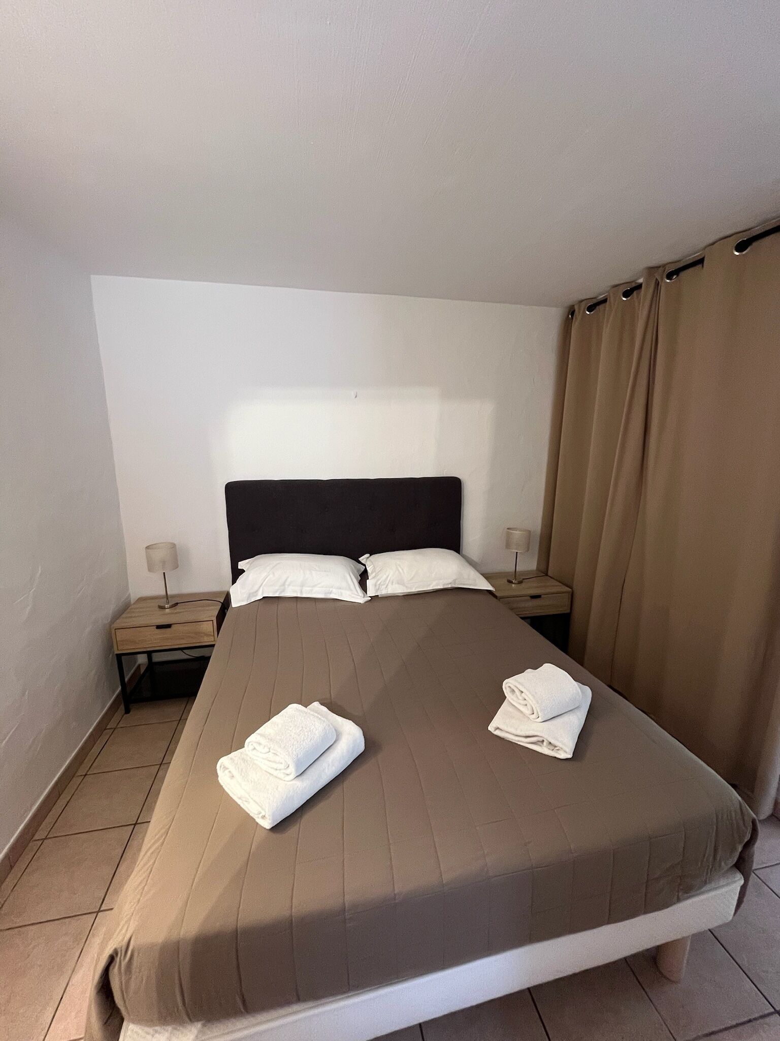 Un lit simple avec une tête de lit, deux oreillers, deux tables de chevet avec des lampes et un rideau.