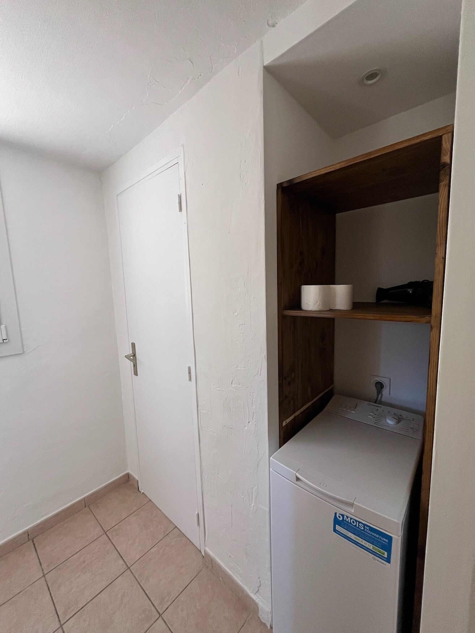 A small, clean room with a white door, a wooden shelf with a white container, and a white appliance with a blue sticker.