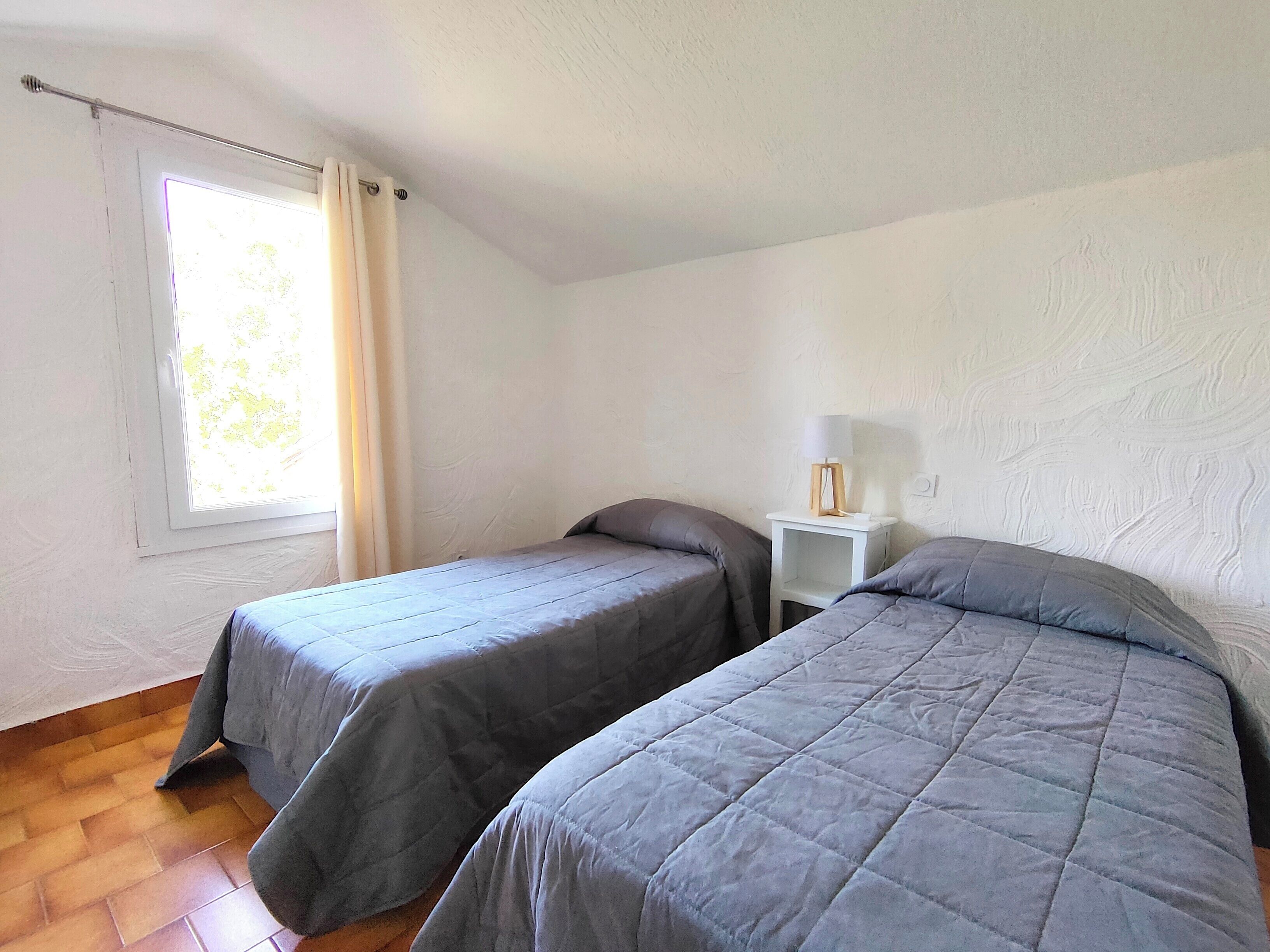 A room with two single beds, a small bedside table with a lamp, a window with curtains, and a wooden floor.