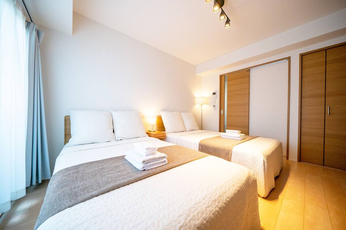 A hotel room with two beds, a wooden wardrobe, a bedside table with a lamp, and a window with curtains.