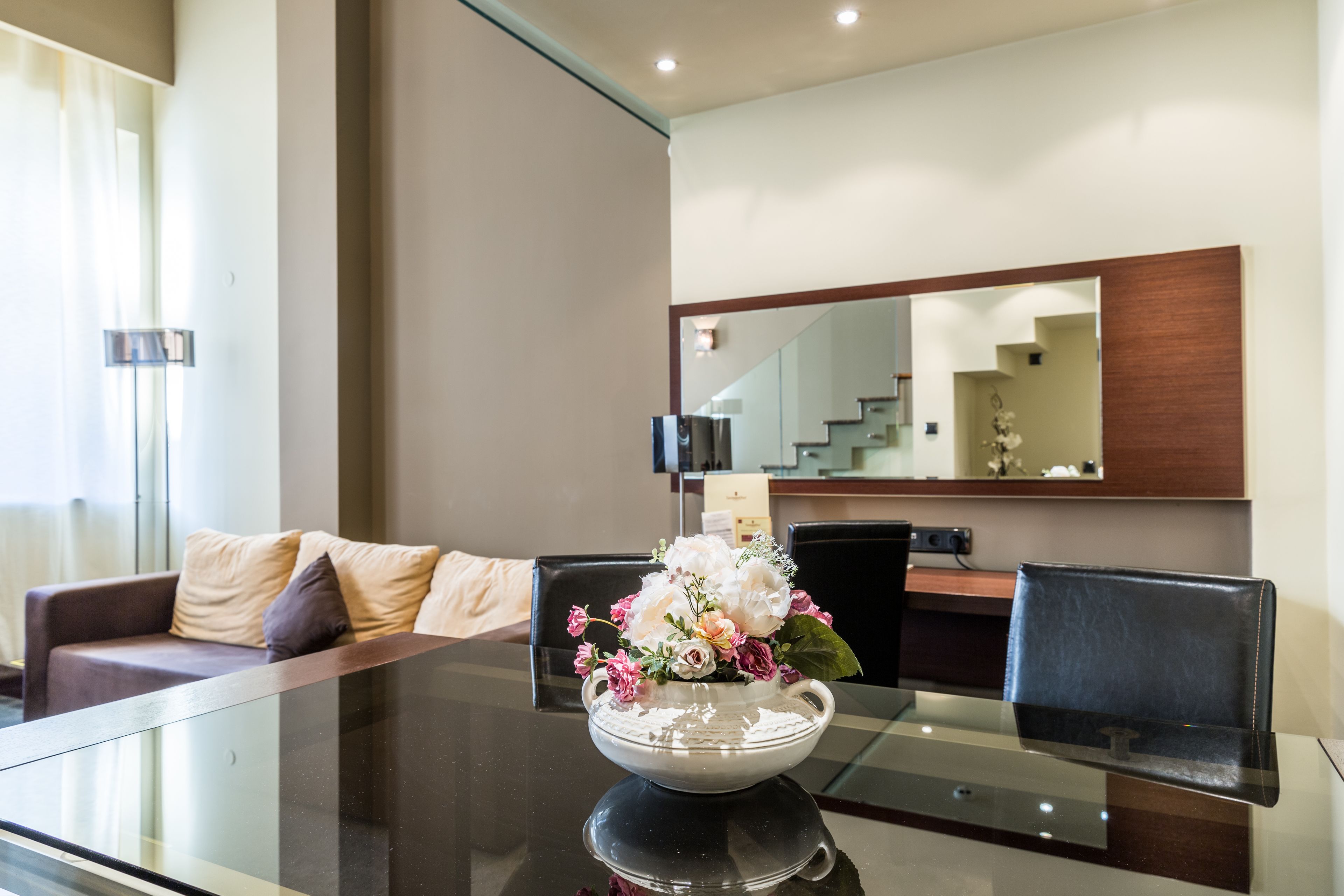 A modern hotel room with a glass dining table, a white vase with flowers, a sofa with cushions, and a large mirror on the wall.
