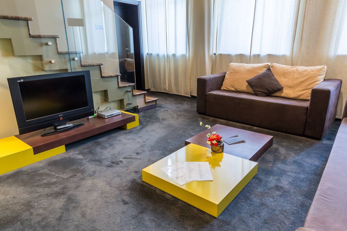 A modern living room with a flat-screen TV, a sofa with cushions, a coffee table with a vase, and a staircase with glass railings.