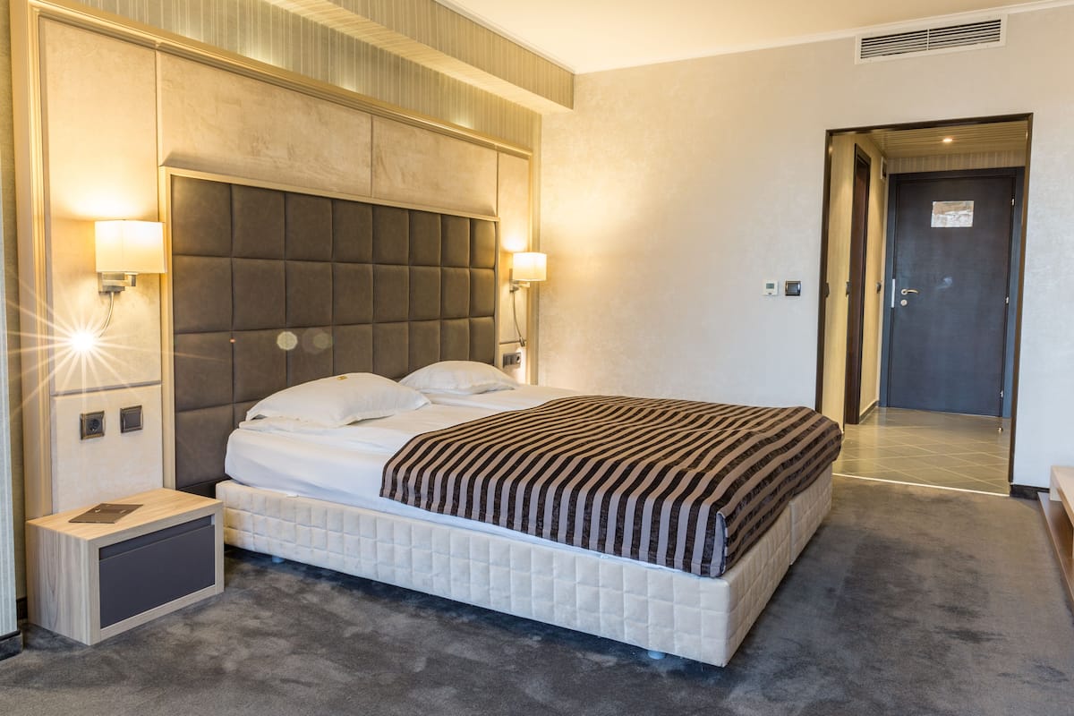 A hotel room with a large bed, two bedside lamps, a nightstand, a headboard, and a door.
