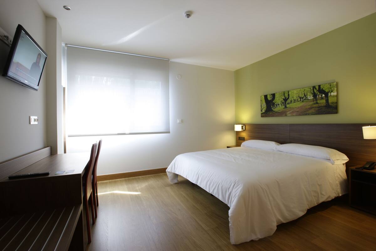Quarto de hotel com cama, escrivaninha e cadeira, televisão, janela com persianas e um quadro na parede.