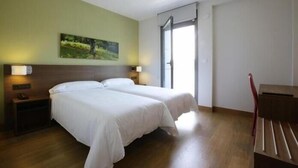 A hotel room with two beds, a nightstand, a desk, and a window with a view of greenery. - Aginaga Hotela (Usurbil)