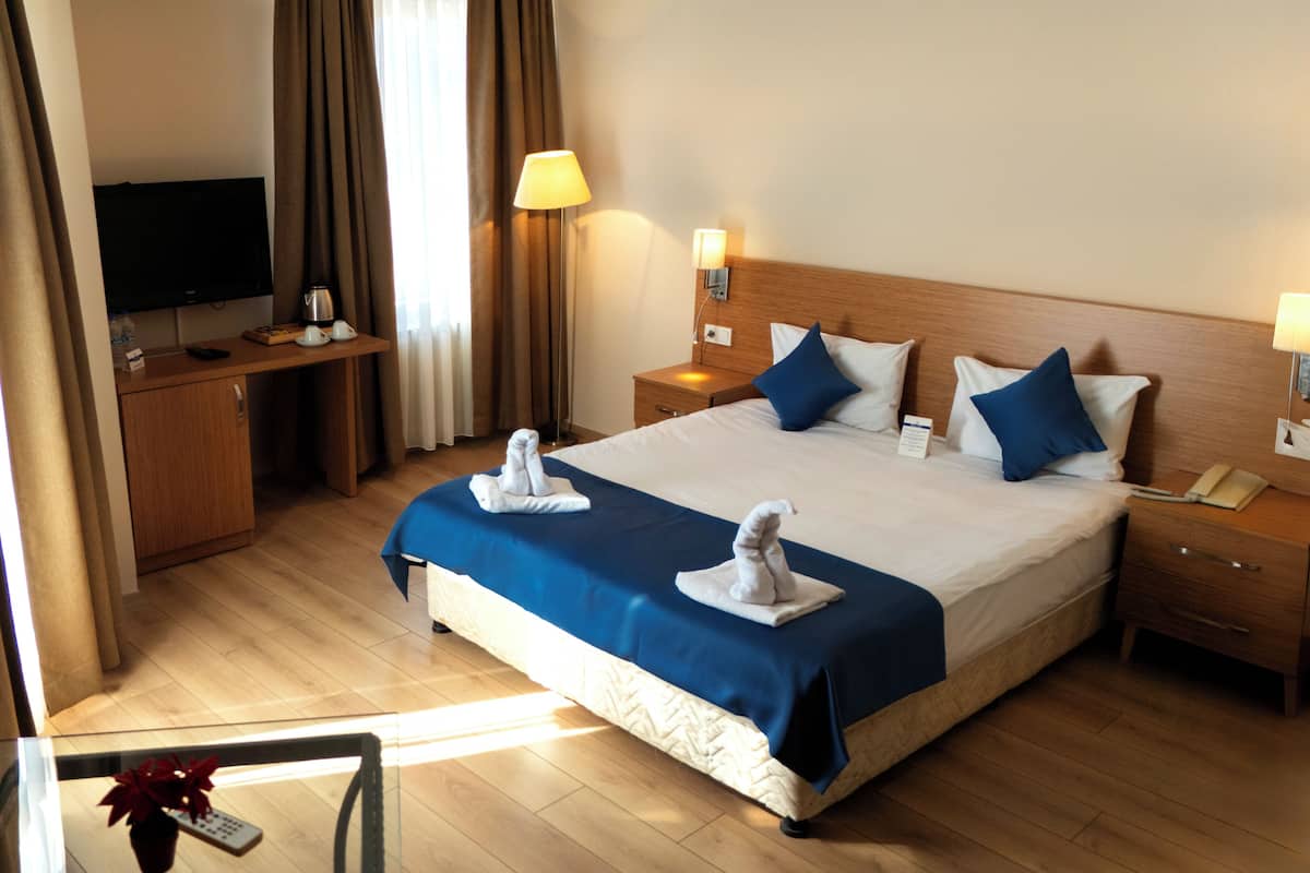 A hotel room with a large bed, two bedside tables, a desk with a flat-screen TV, a lamp, and a remote control on a glass table.
