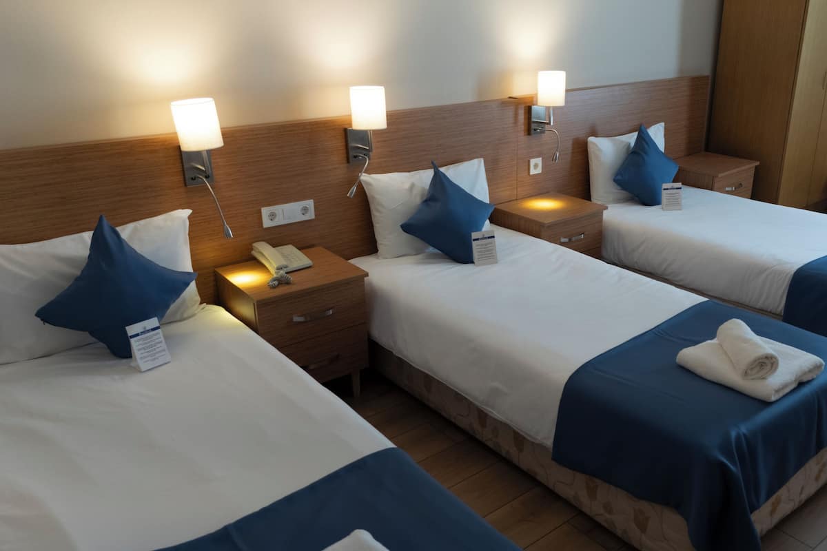 A hotel room with three beds, each with a blue bedspread and white pillows. There are bedside lamps, a wooden headboard, and a small wooden table with a chair.