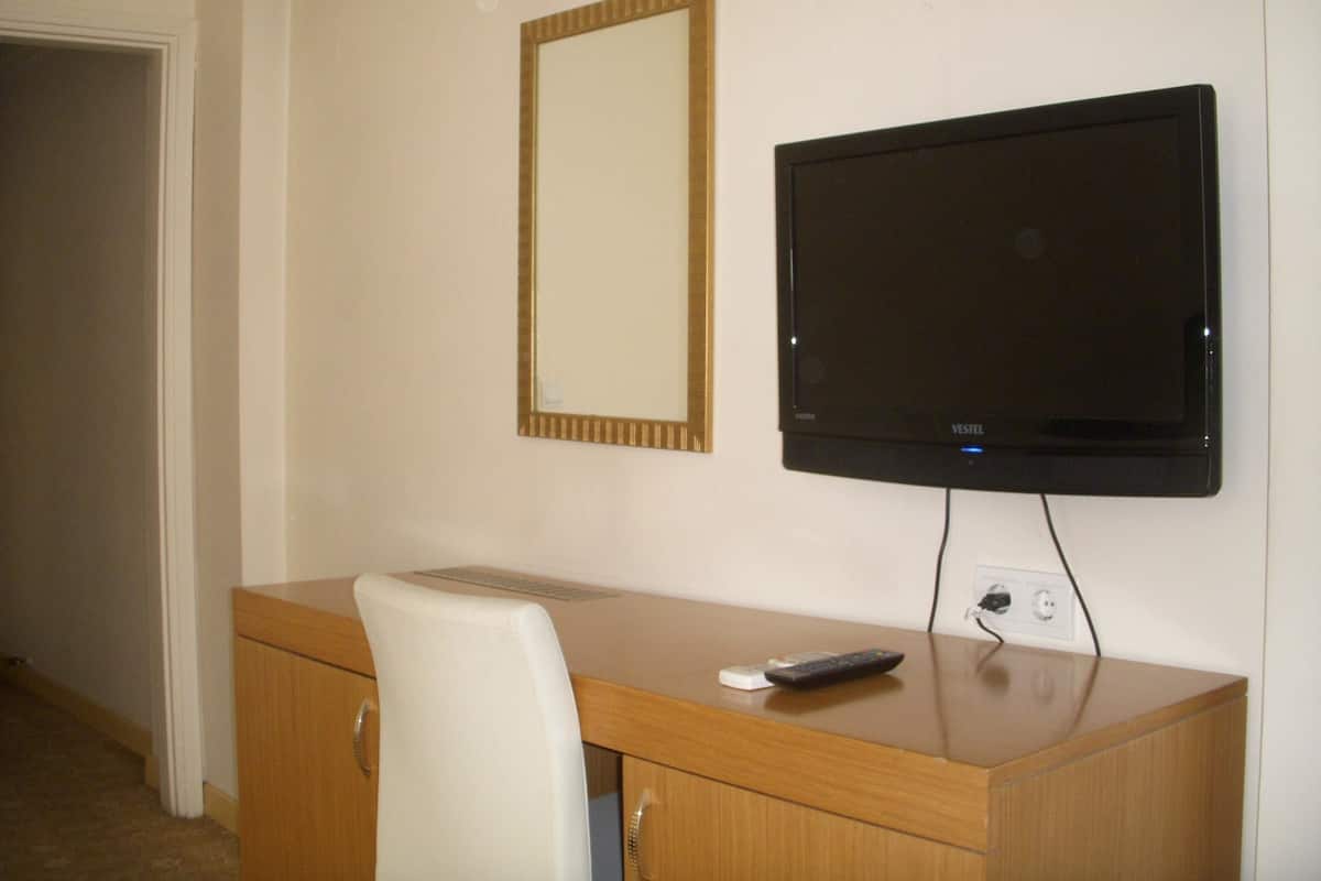 A hotel room with a wooden desk, a white chair, a flat-screen TV mounted on the wall, and a mirror above the desk.