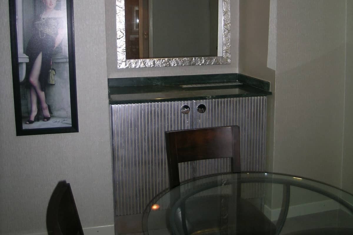 A dining area with a glass table, a wooden chair, a framed picture on the wall, and a mirror above the countertop.