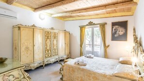 Een slaapkamer met een houten plafond, een groot versierd kastje, een bed met een bloemenbedekking en een schilderij aan de muur.