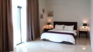 A hotel room with a large bed, two bedside tables with lamps, a window with curtains, and artwork on the walls. - Balinese House (Malacca City)