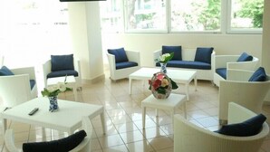 Un área de estar luminosa y moderna con mobiliario blanco y cojines azules, que cuenta con un televisor de pantalla plana y grandes ventanales con vistas al exterior.
