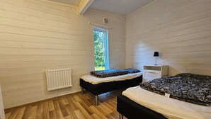 A room with two beds, a radiator, a window with a view of greenery, and a white nightstand with a lamp.