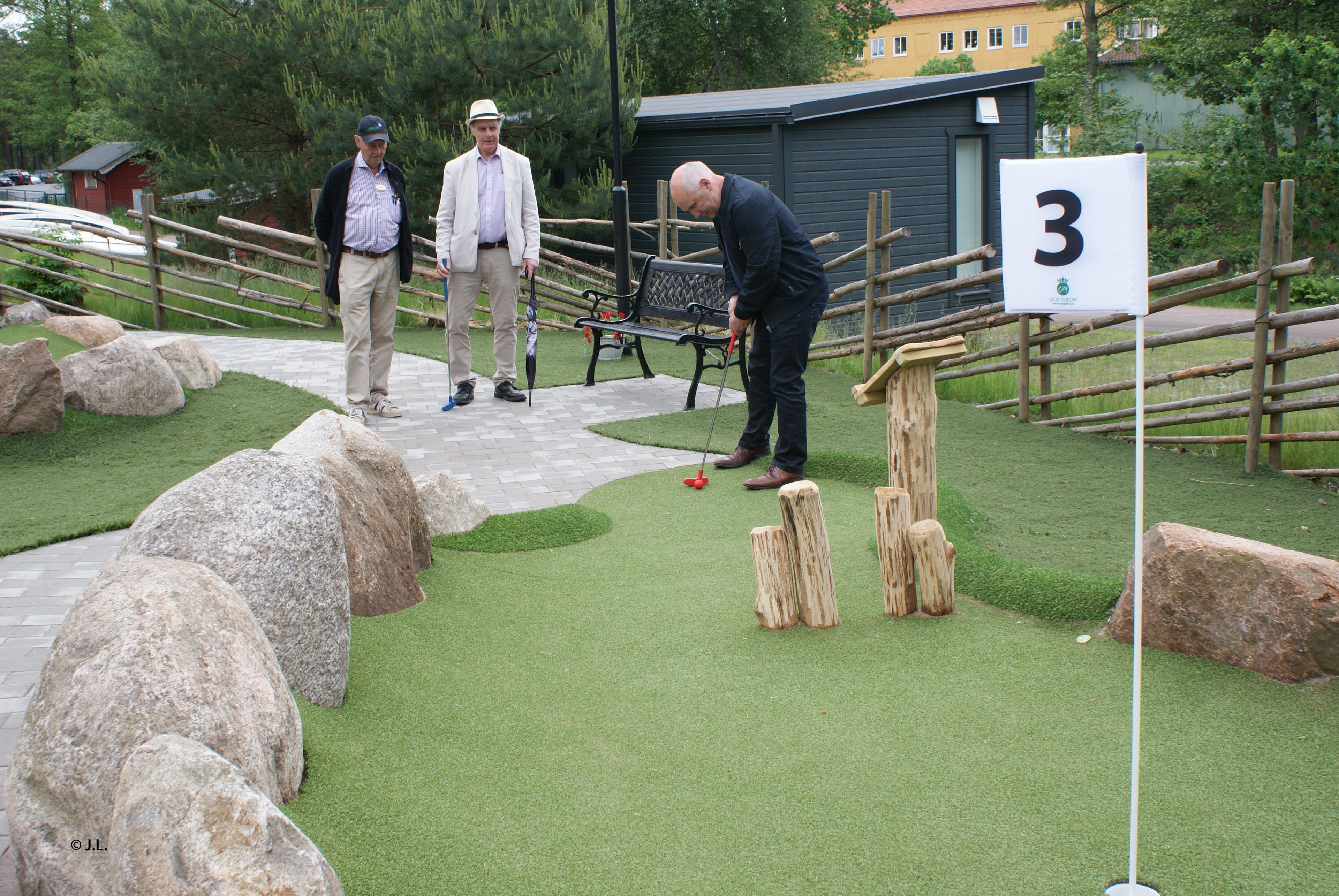 Un campo de mini golf con una valla de madera, un letrero con el número 3 y una banca.