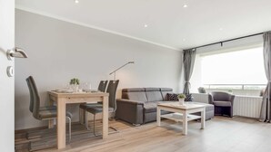 A modern living room with a sofa, a dining table with chairs, a coffee table, and a large window with curtains. - Apartment 93 (ID 082), Germany (Sankt Peter-Ording)