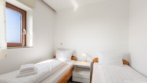 Quarto pequeno e limpo com duas camas de solteiro, mesa de cabeceira com abajur e janela com persianas.