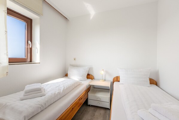 A small, clean room with two single beds, a nightstand with a lamp, and a window with blinds.