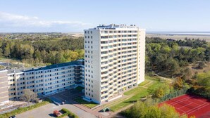 Um edifício alto com vários andares, rodeado por uma área arborizada e uma quadra esportiva.