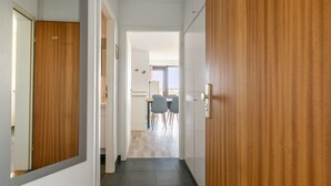 Corredor com porta de madeira, parede branca e chão de azulejos, conduzindo a uma sala de jantar com mesa e cadeiras.