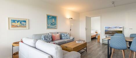 A modern living room with a grey sofa, a wooden coffee table, and a dining area with blue chairs.