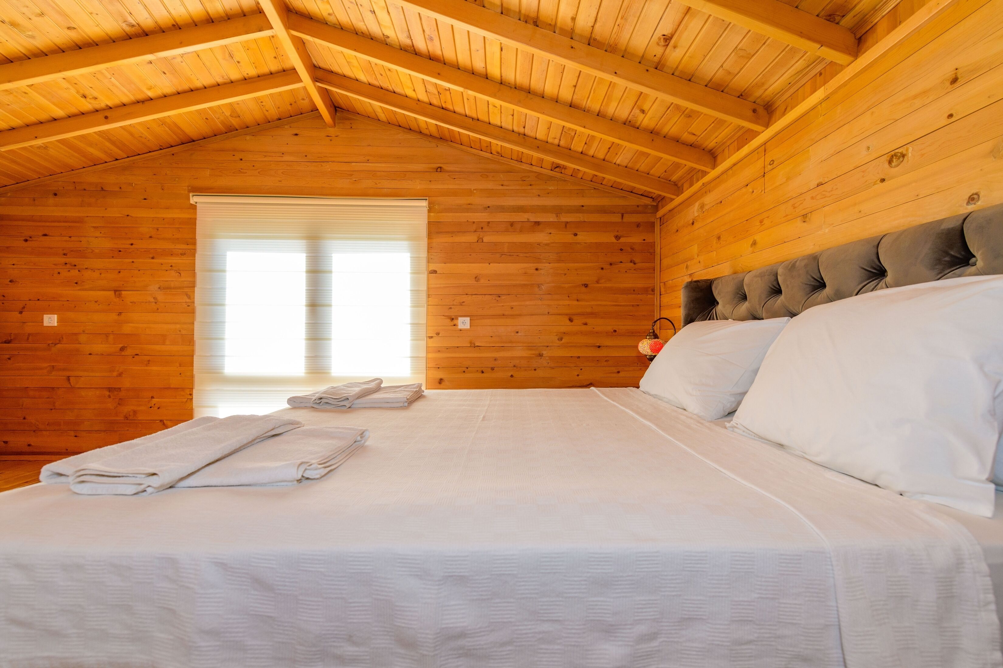 Une pièce de cabane en bois comprenant un lit, une fenêtre avec des stores et un plafond avec des poutres en bois.