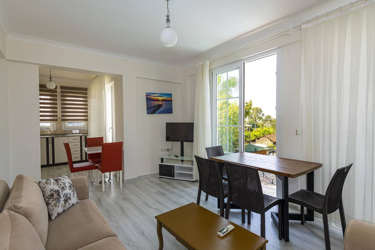 A modern living room with a sofa, a wooden coffee table, and a dining area with a table and chairs. There is a TV and a painting on the wall.