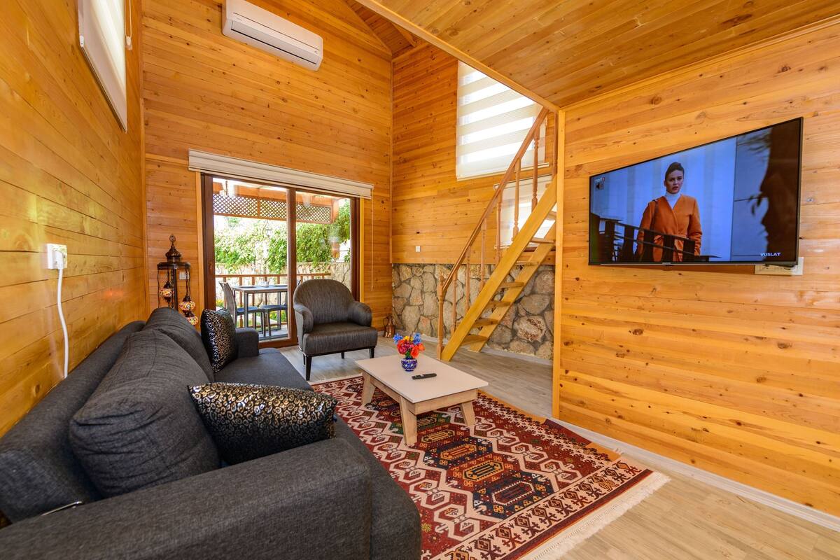 A wooden cabin interior with a sofa, armchairs, a coffee table, and a TV on the wall.