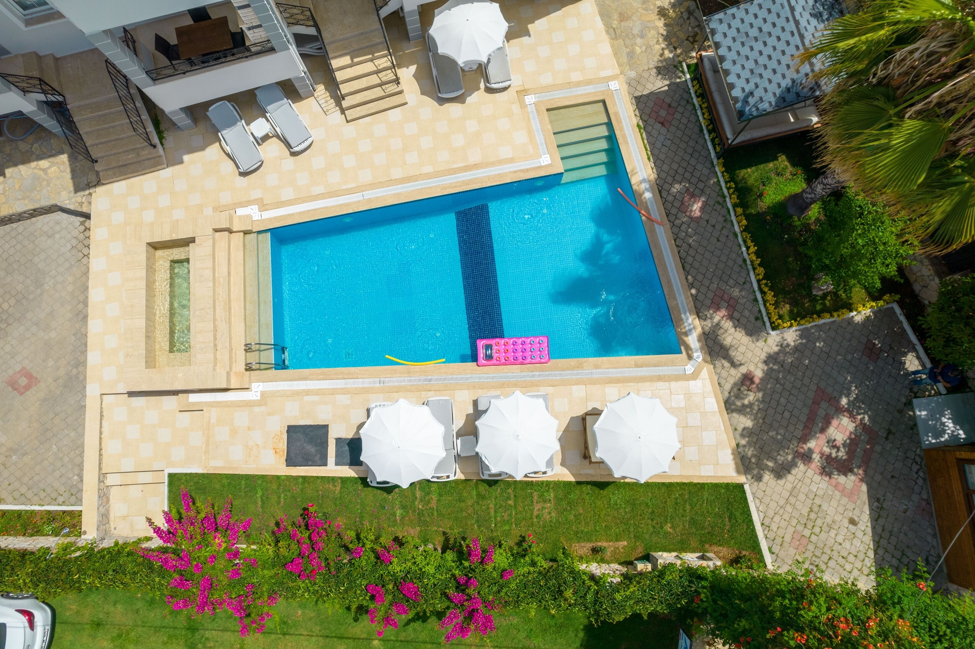 An aerial view of a swimming pool area with a blue pool, white lounge chairs, and a small garden with pink flowers.