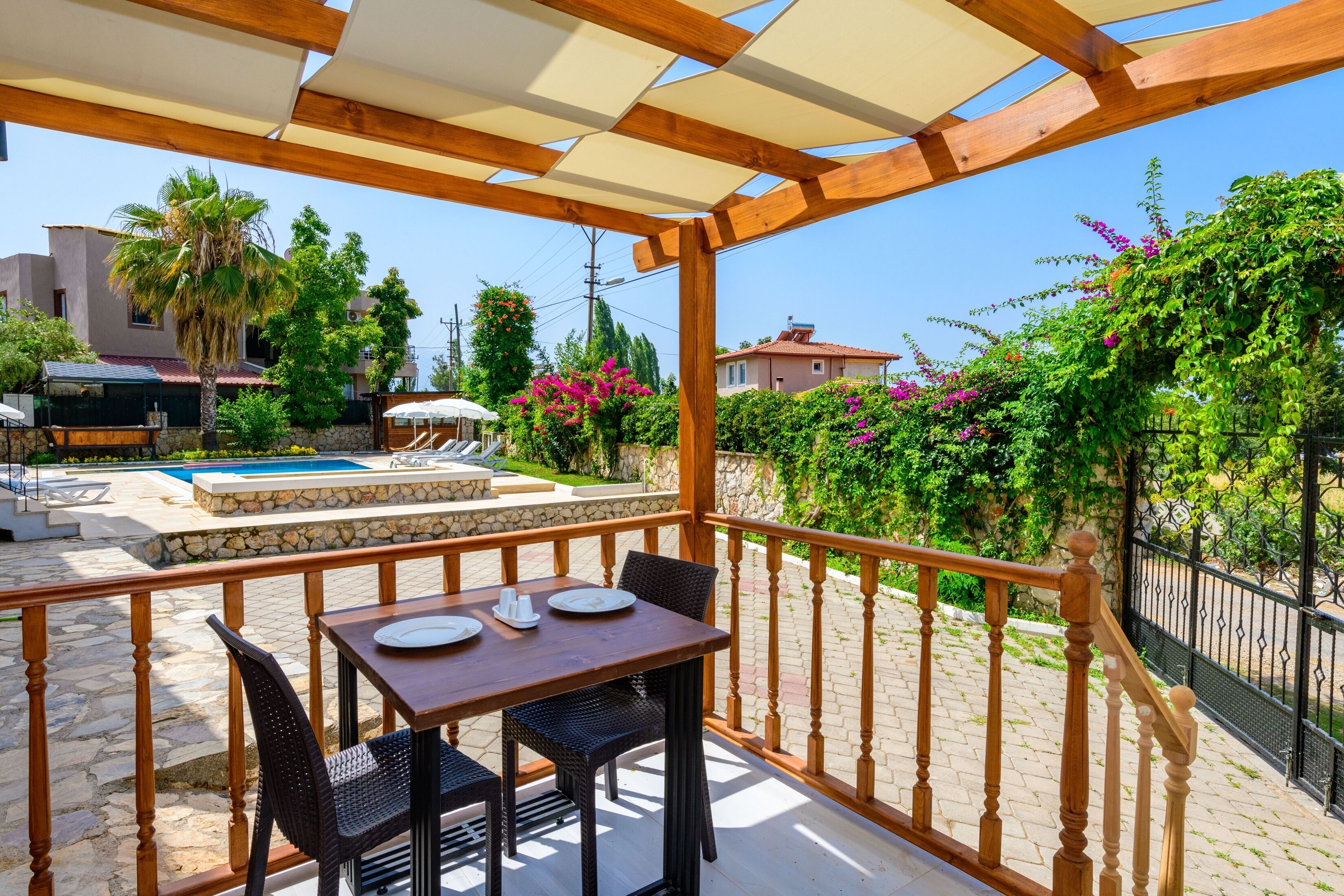 A covered outdoor area with a wooden table and chairs, a stone pool, and a view of the surrounding greenery and buildings.