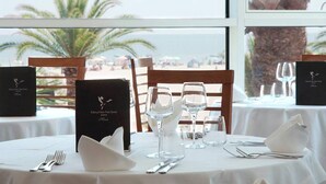 Mesa lista para comer con un menú, vasos y cubiertos, con vista a una playa con palmeras.