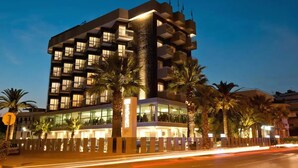Un hotel de varios pisos con balcones, fachada de vidrio y palmeras en la entrada.