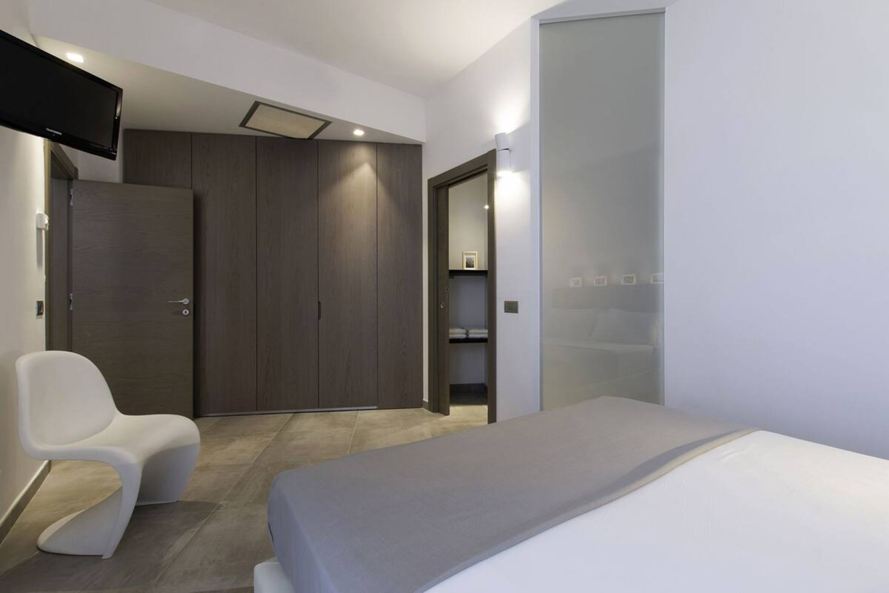 Una habitación de hotel moderna con una cama, una silla blanca, un armario y un televisor montado en la pared.