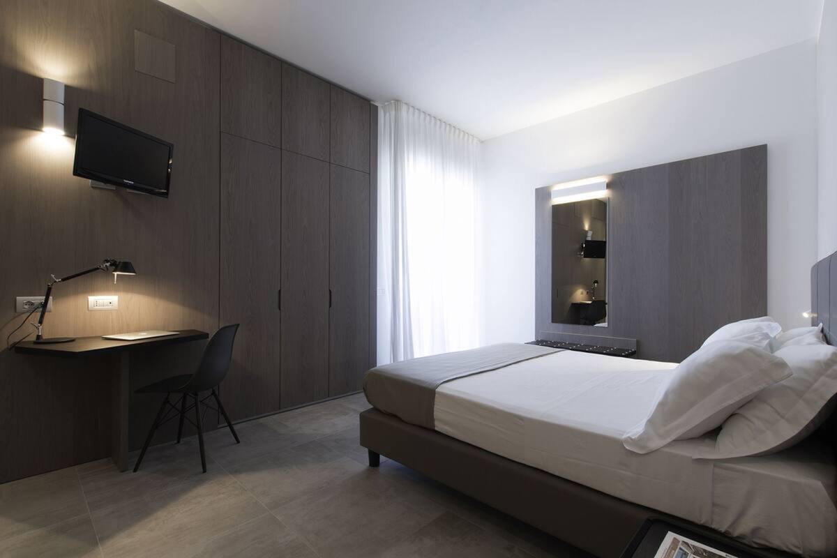 A modern hotel room with a bed, desk, chair, and a TV mounted on the wall.