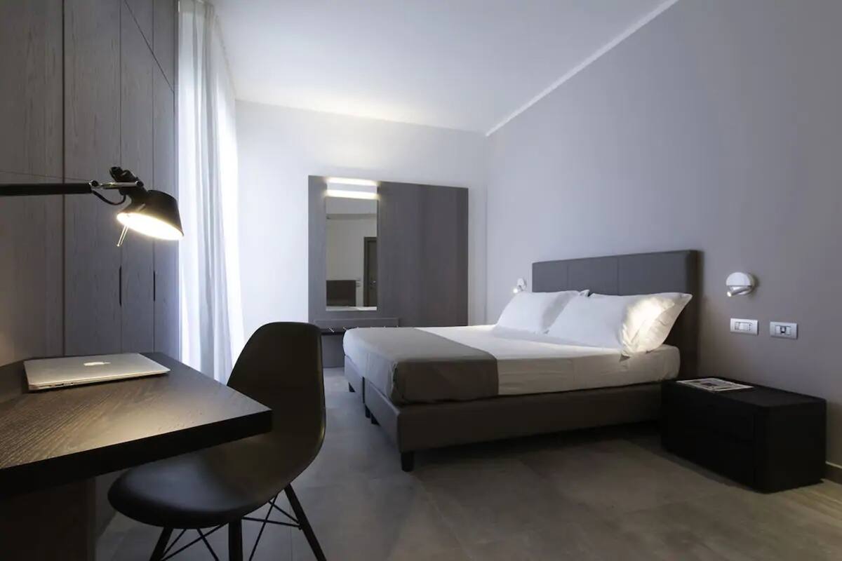 A modern hotel room with a bed, a desk, and a chair. There is a lamp on the desk and a small table beside the bed.