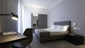 Une chambre d’hôtel moderne, équipée d’un lit, d’un bureau et d’une chaise. On trouve une lampe sur le bureau et une petite table à côté du lit.
